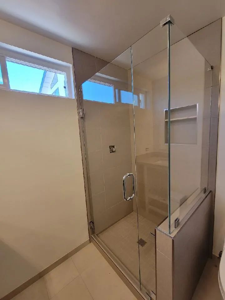 Glass shower enclosure in a beige-tiled bathroom with a window above.
