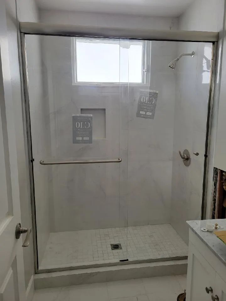 Small white walk-in shower with glass doors, grab bar, window, and tiled floor.