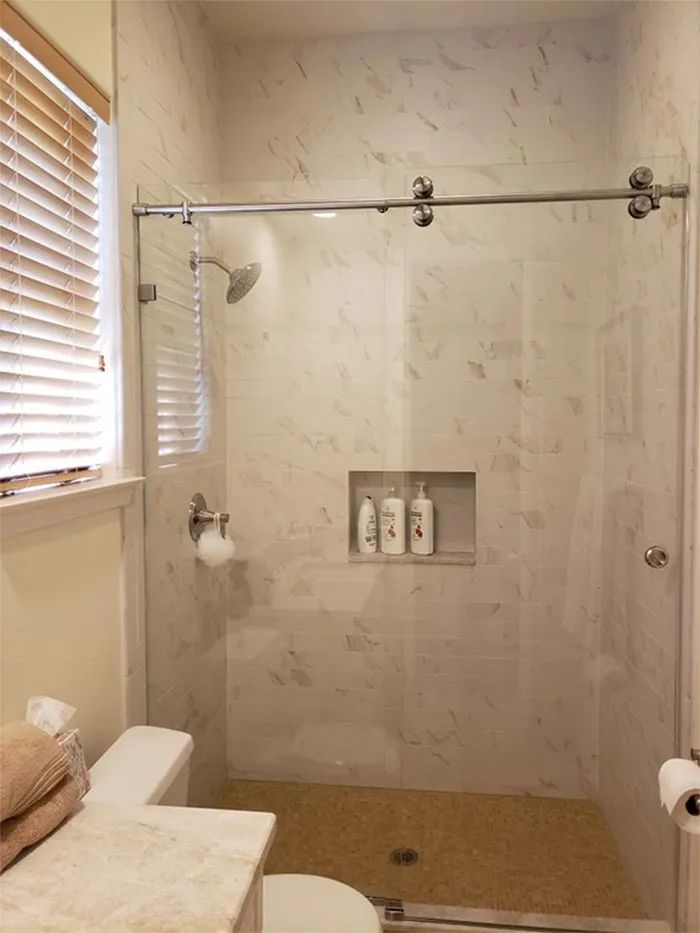 Glass walk-in shower with beige tile, built-in shelf, and sliding door hardware next to a window.
