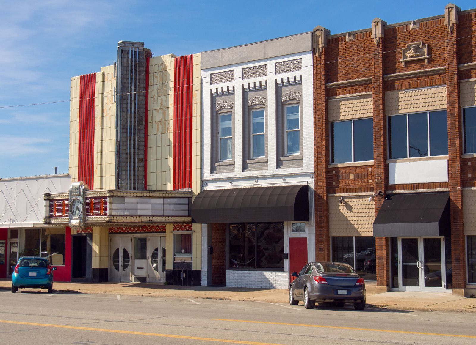 Row of historic buildings on a street, including an art deco theater and shops, cars parked in front.