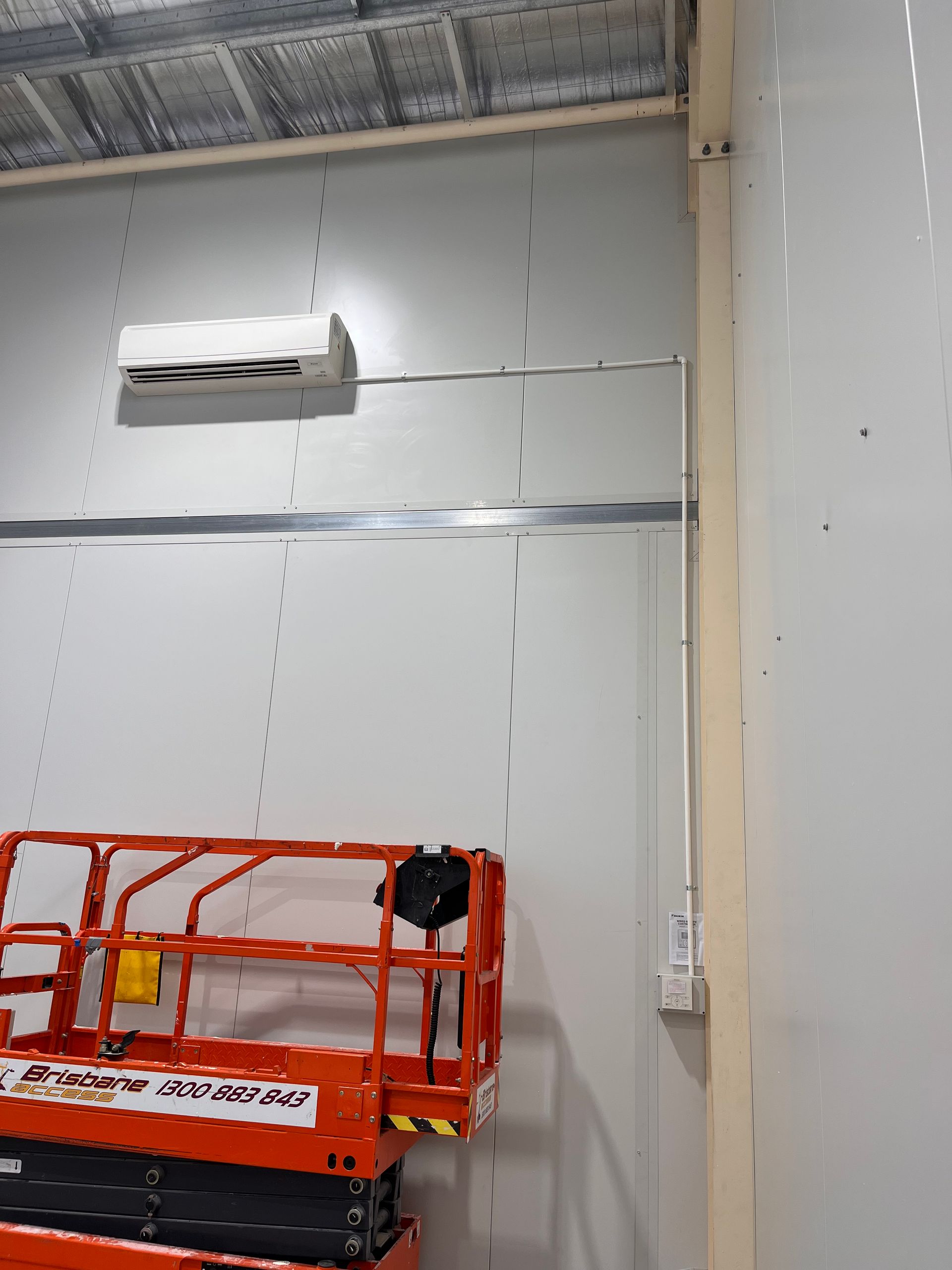 White industrial wall with mounted AC unit, electrical outlet, and orange scissor lift in a warehouse.