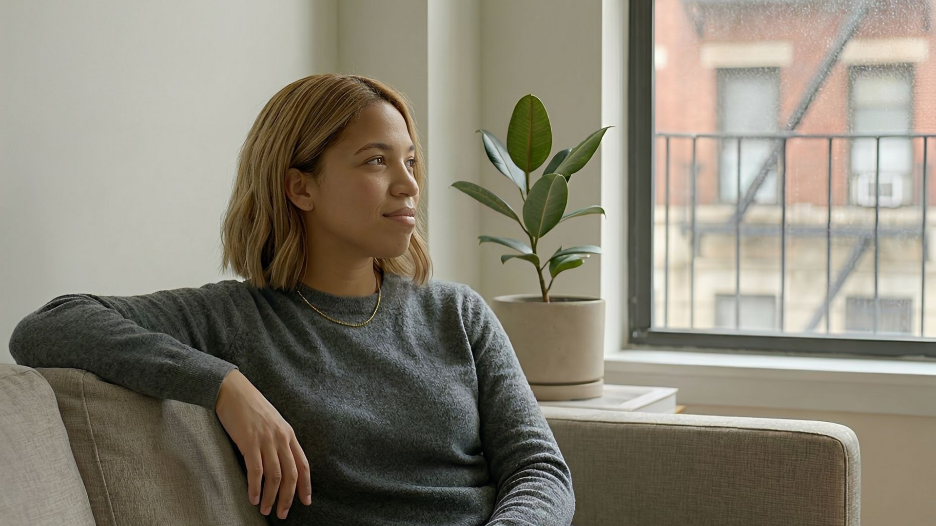 Woman sitting on a couch, looking out window, houseplant beside her.