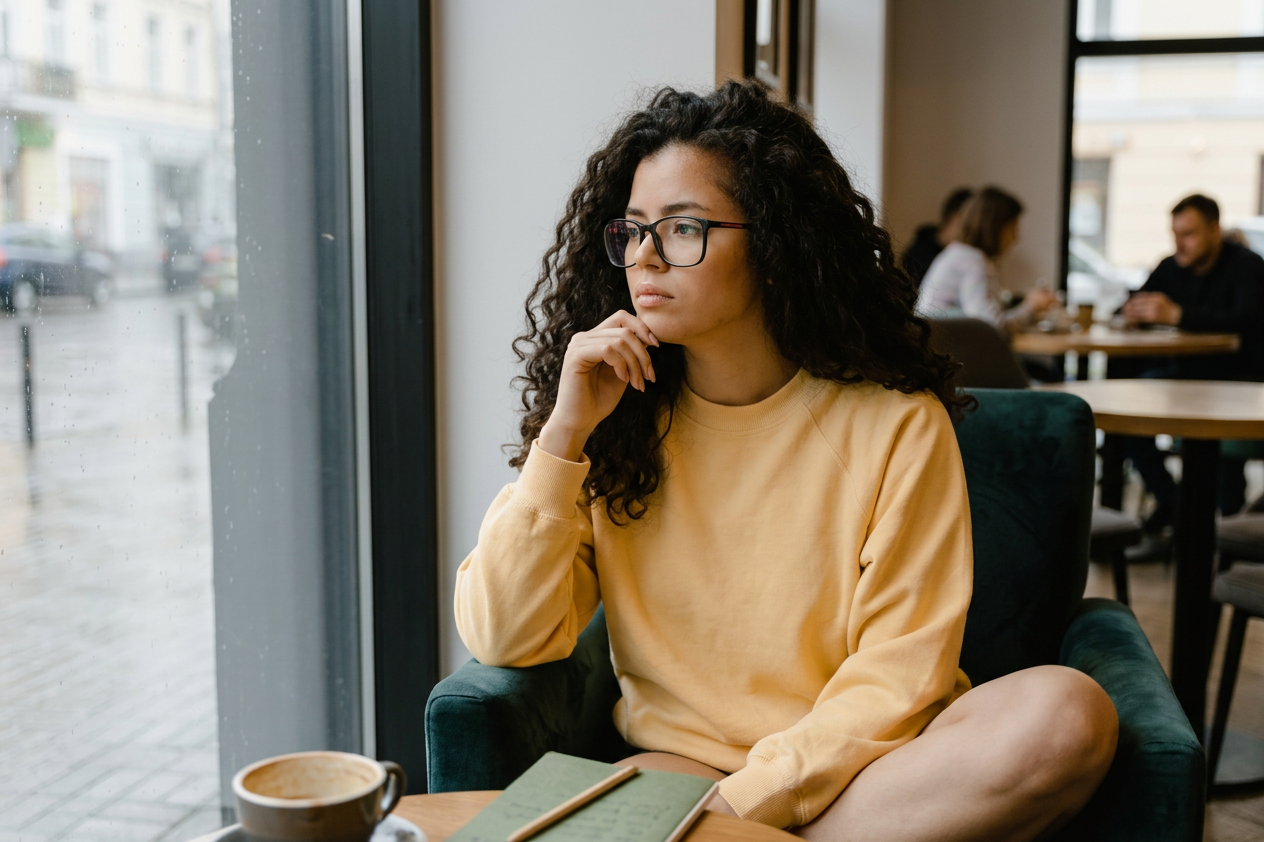 Woman in a mustard sweater sitting by a window in a cafe, looking thoughtful