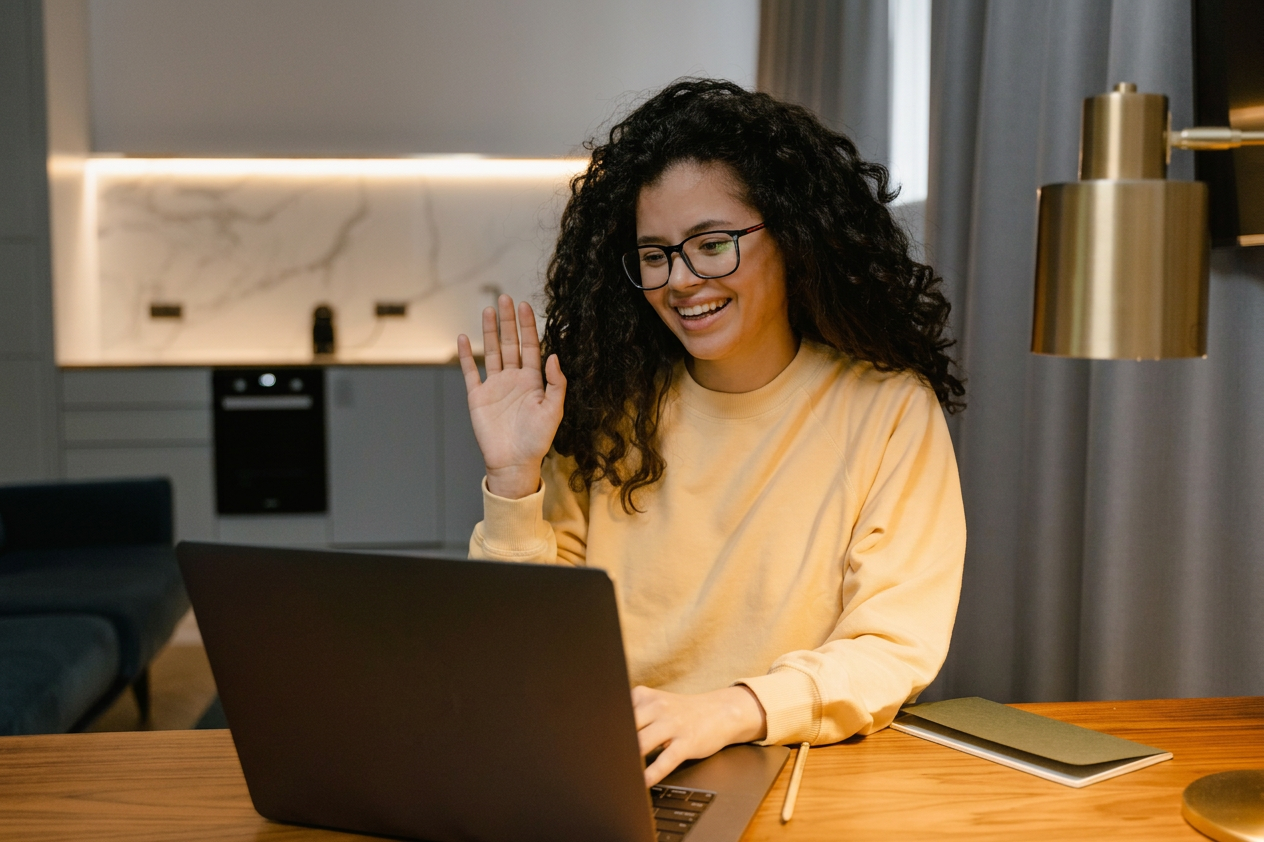 Woman smiling and waving from behind a laptop at a wooden desk in a modern kitchen workspace
