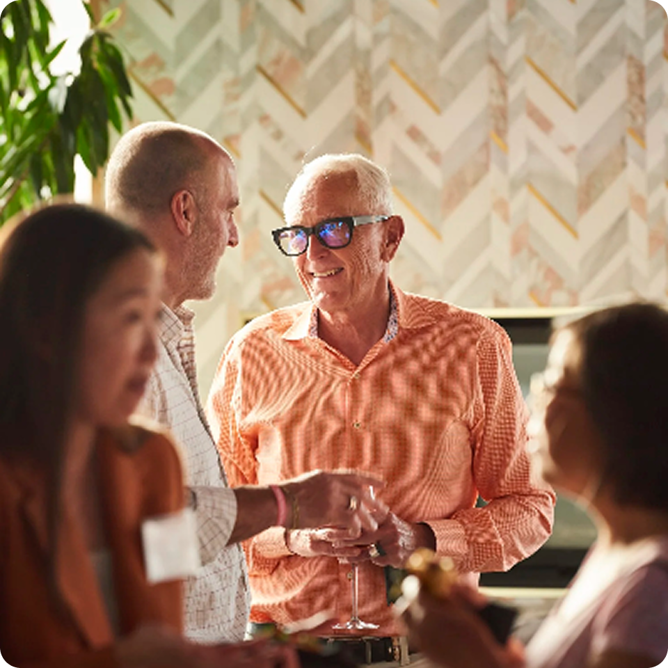 People socializing at an event, man in orange shirt smiling, holding a drink.
