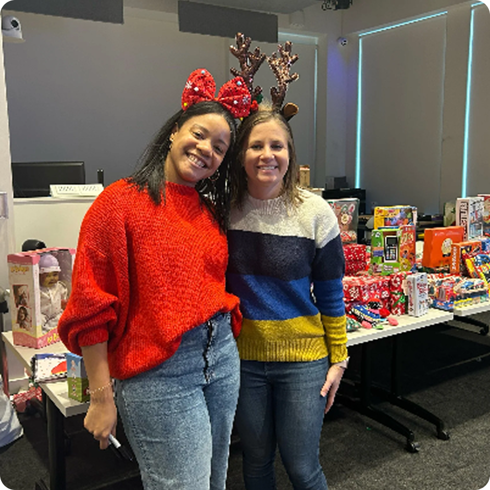 Two people wearing festive headbands pose by a table filled with gifts.