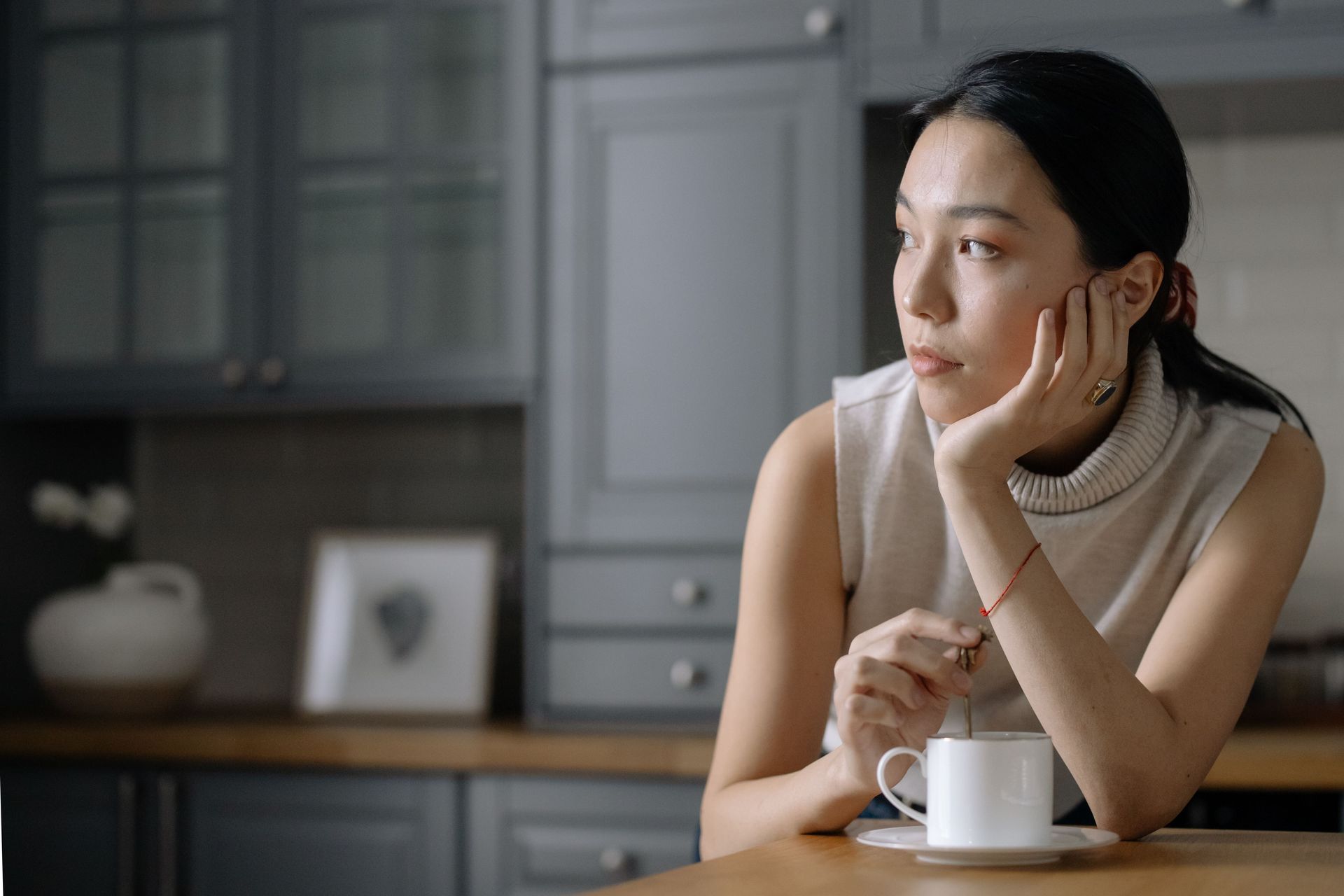 Person resting chin on hand beside a white mug in a gray kitchen, gazing out the window.