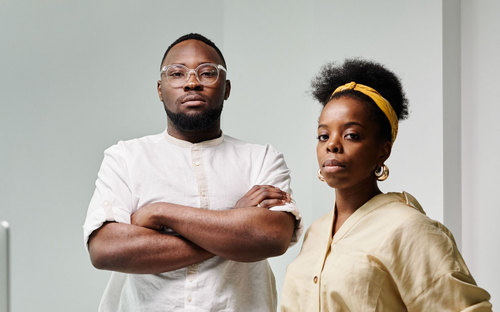 Two people standing against a light gray wall, arms crossed, wearing neutral-toned shirts.