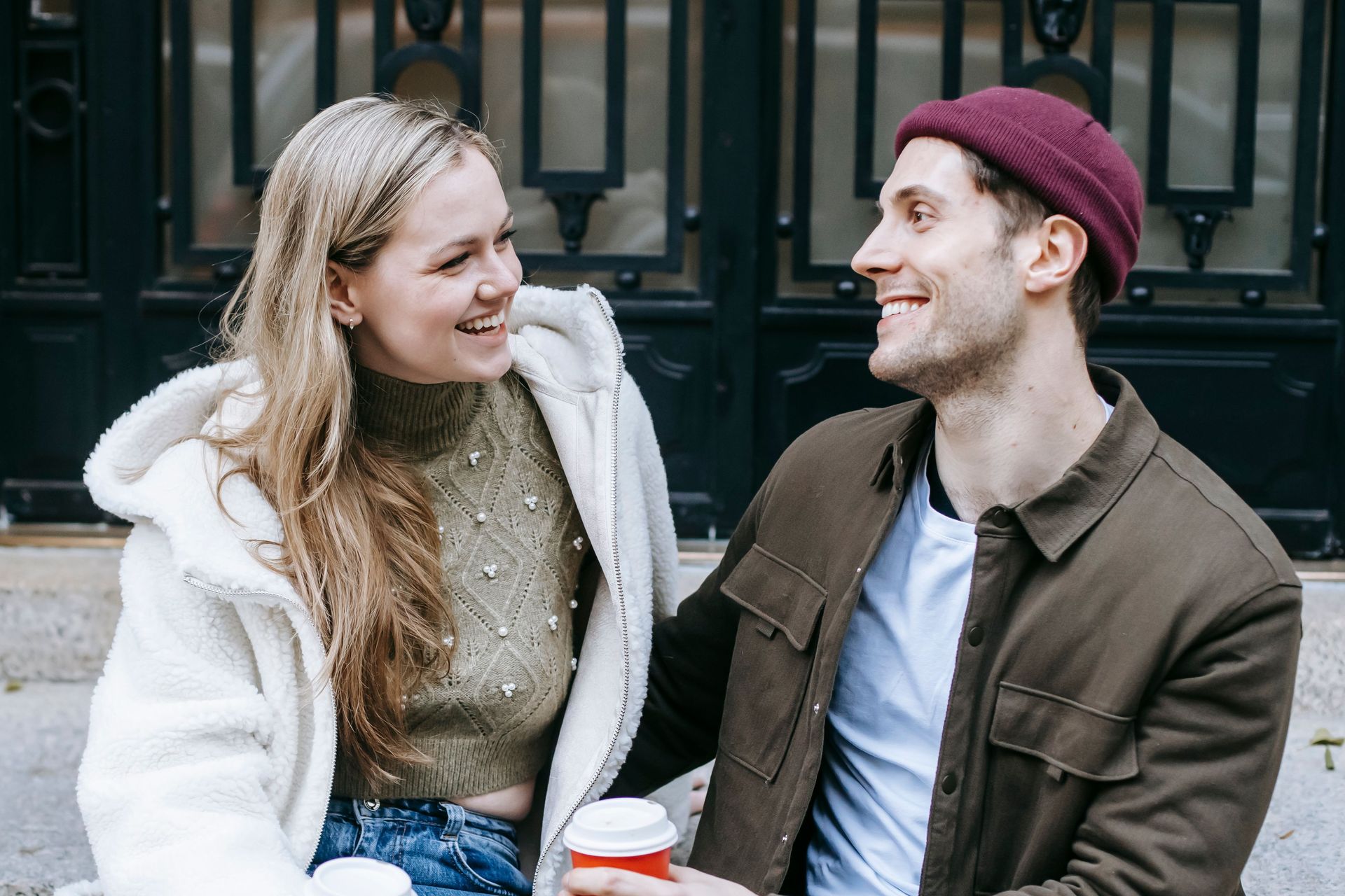 Two people smiling and talking on a bench, one in a white coat and one in a maroon beanie, holding coffee cups