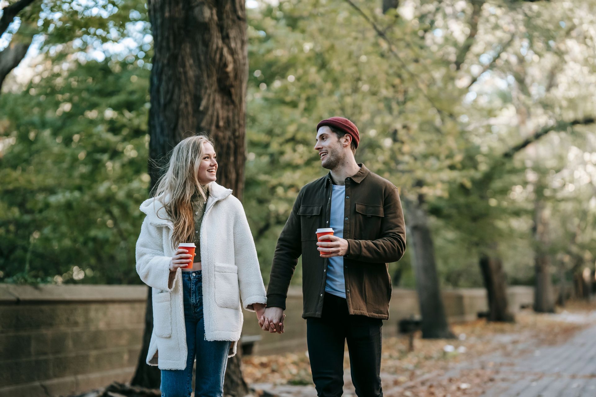 Two people walking hand in hand with coffee cups in a leafy park, smiling at each other