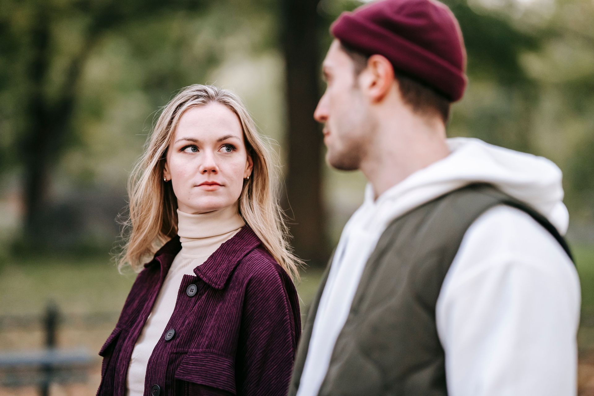 Two people outdoors; a woman in a burgundy jacket looks at a man in a maroon beanie and gray hoodie.