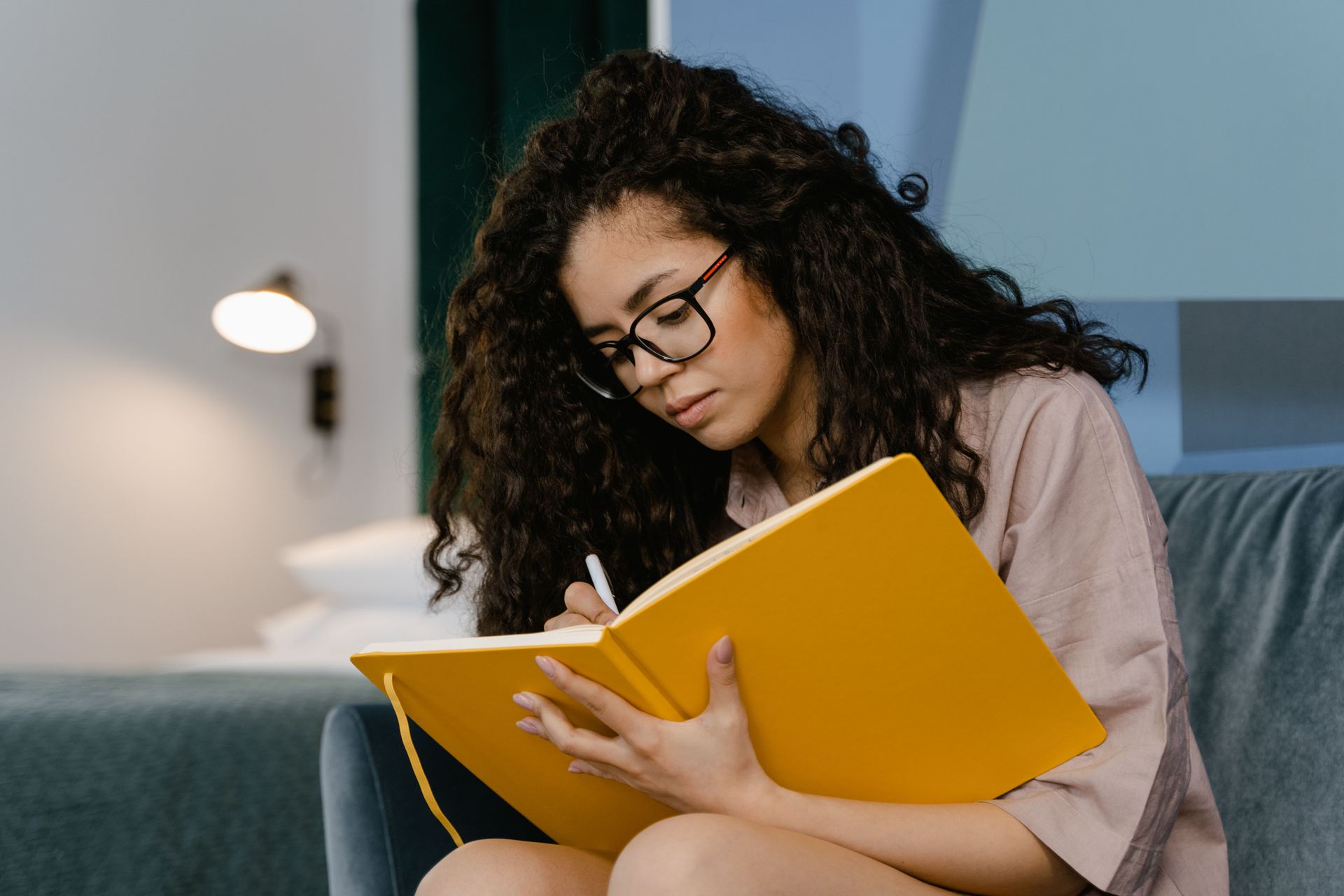 Person writing in a yellow notebook on a couch beside a lamp.