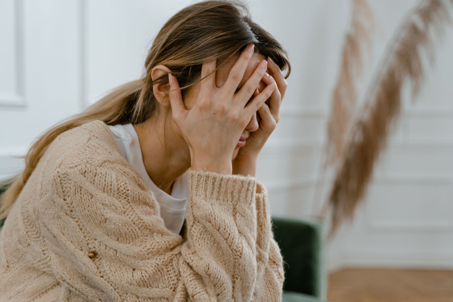 Woman in a beige sweater covering her face with her hands, sitting indoors looking distressed