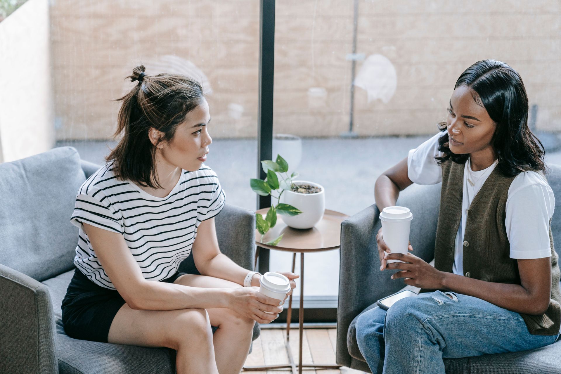 Two people talking on armchairs in a bright living room, one holding a cup, with a small table and plant between them