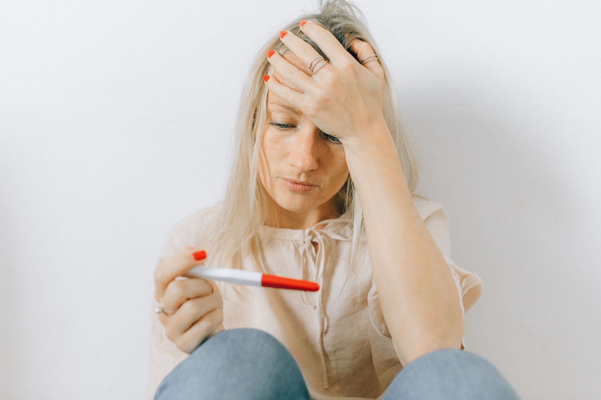 Woman holding a pregnancy test and touching her forehead, looking stressed against a plain white background