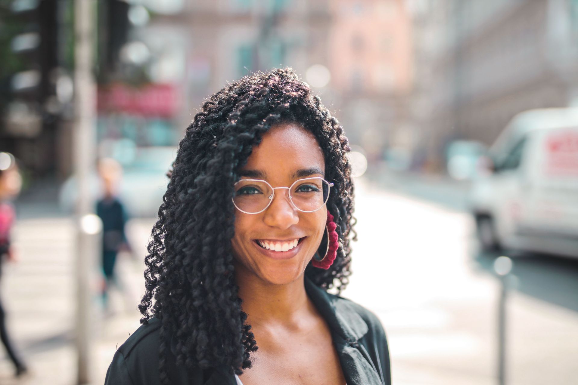 Smiling woman with glasses standing on a city street, with blurred cars and buildings in the background