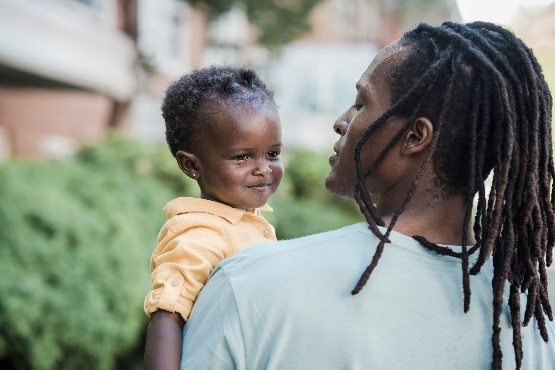 Adult holding a smiling child outdoors on a sidewalk, both facing each other