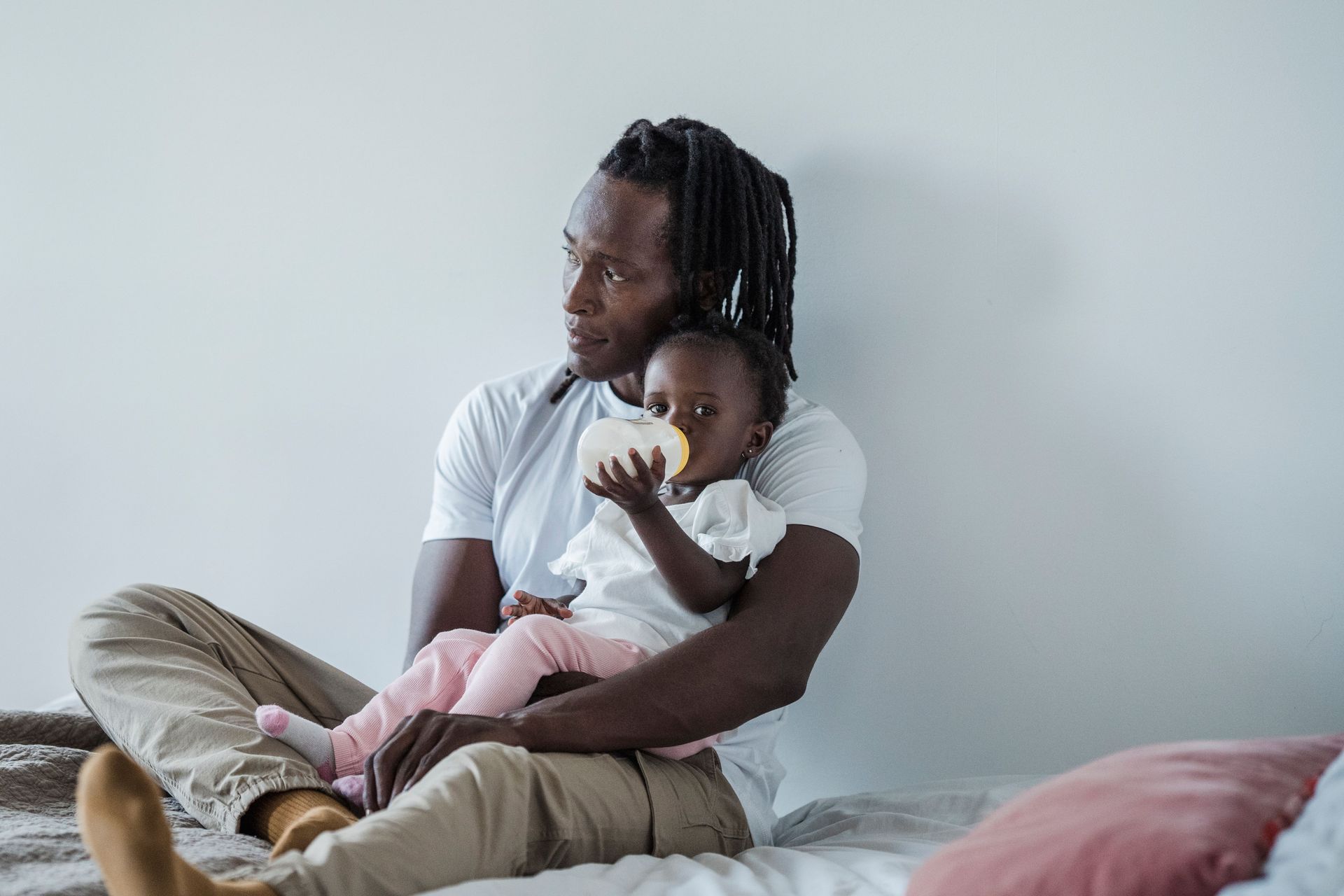 Adult holding a child on a bed in a softly lit bedroom, both wearing white shirts and beige pants.