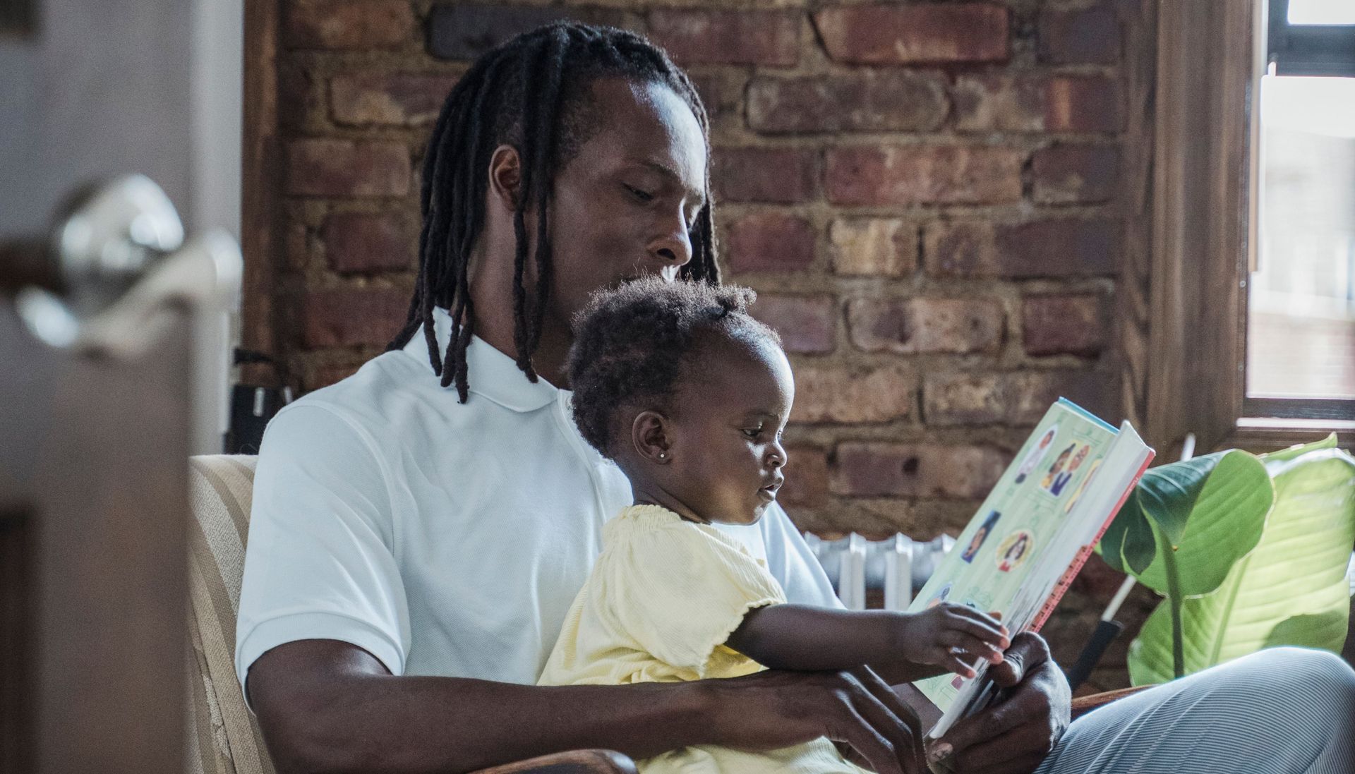Adult and child reading a book together on a couch in a brick-walled room