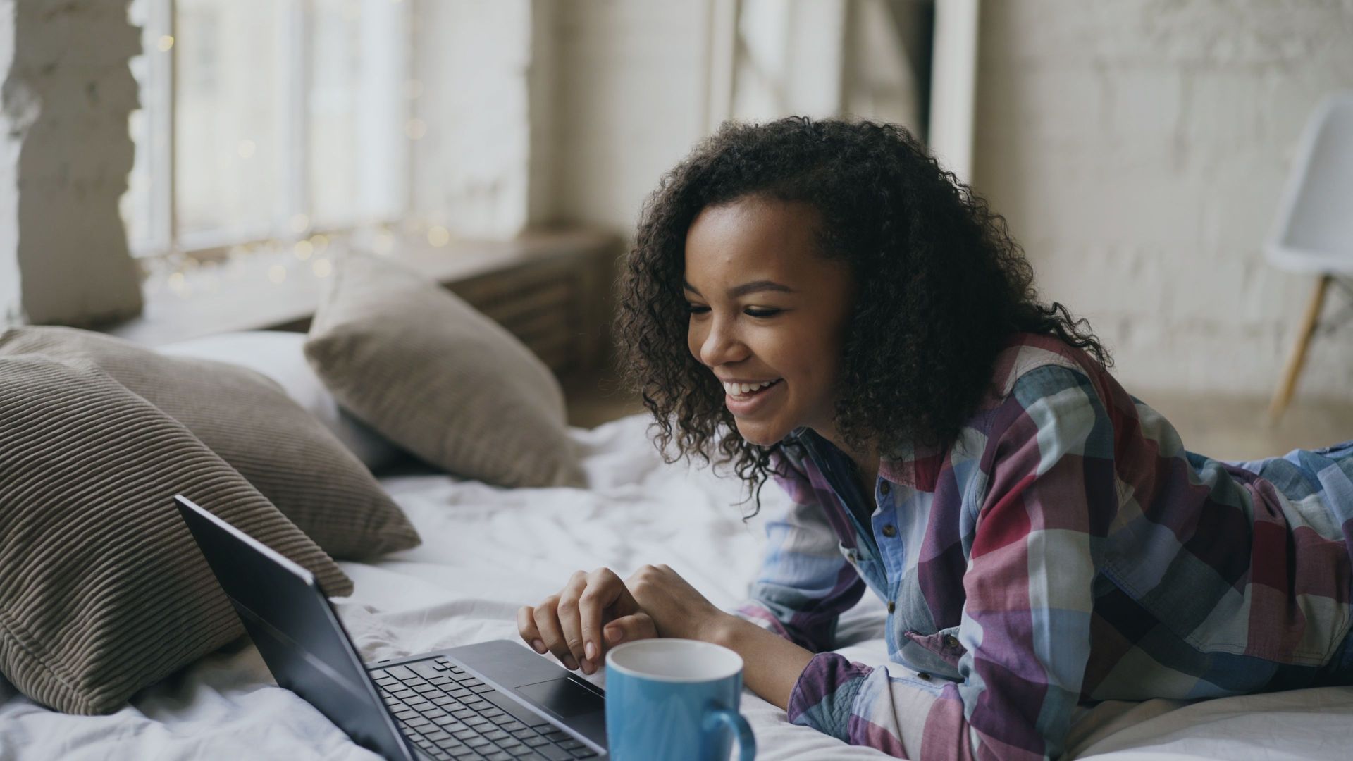 Woman smiling at a laptop on a bed with a blue mug in a bright bedroom