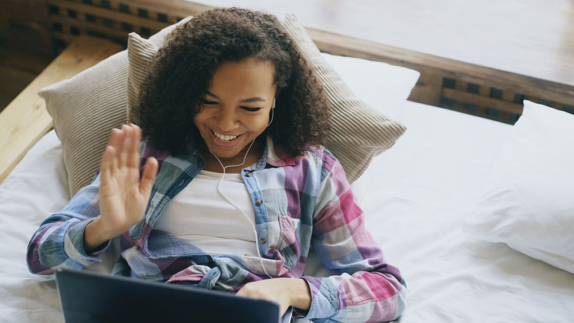 Person smiling and waving while using a laptop on a bed with pillows and a plaid shirt
