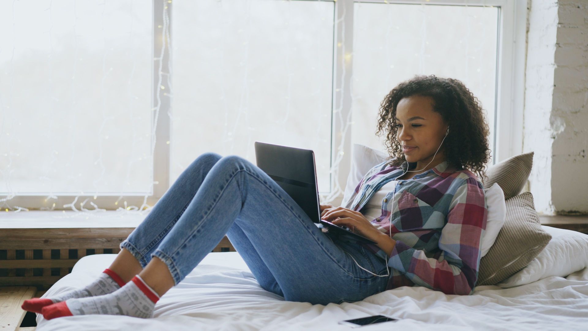Person lounging on a bed with a laptop, wearing a plaid shirt and jeans in a bright room