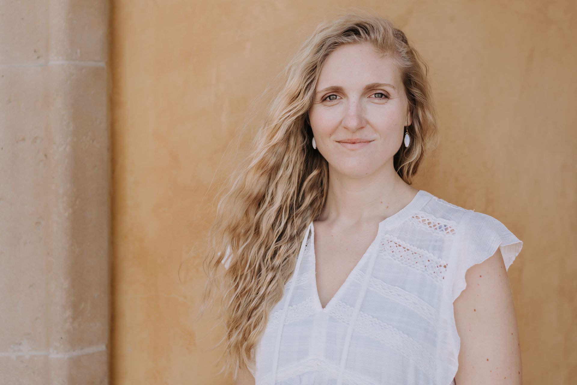 Woman with long wavy blonde hair in a white blouse standing against a tan wall, facing the camera.