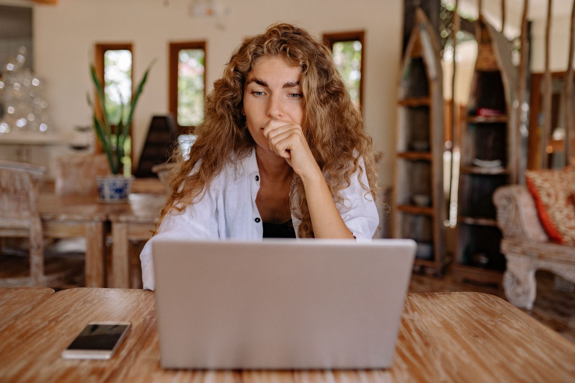 Person with curly hair working on a laptop at a wooden table in a bright room