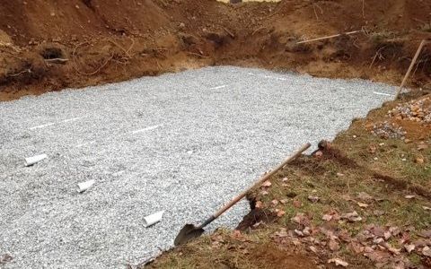 Fosse remplie de gravier dans la terre excavée, pelle appuyée contre le bord, avec des repères blancs.