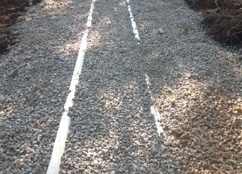 Allée de gravier traversée par des tuyaux en PVC blanc, probablement pour le drainage.