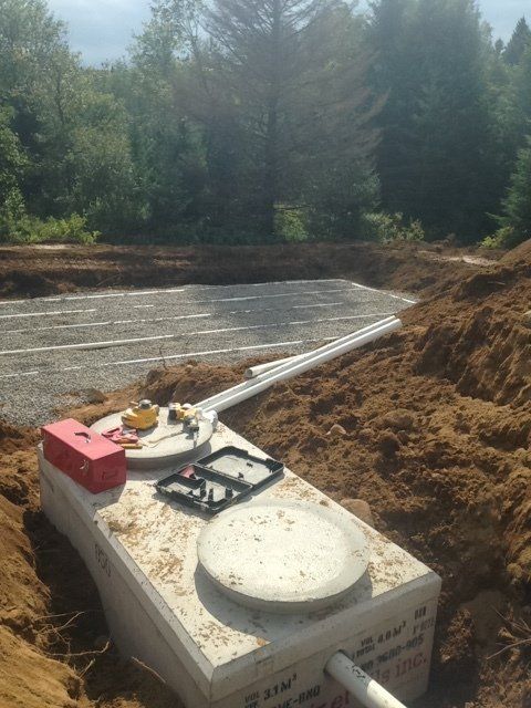 Installation d'une fosse septique en cours ; fosse en béton avec tuyauterie, entourée d'un champ de terre et de gravier. Arrière-plan forestier.