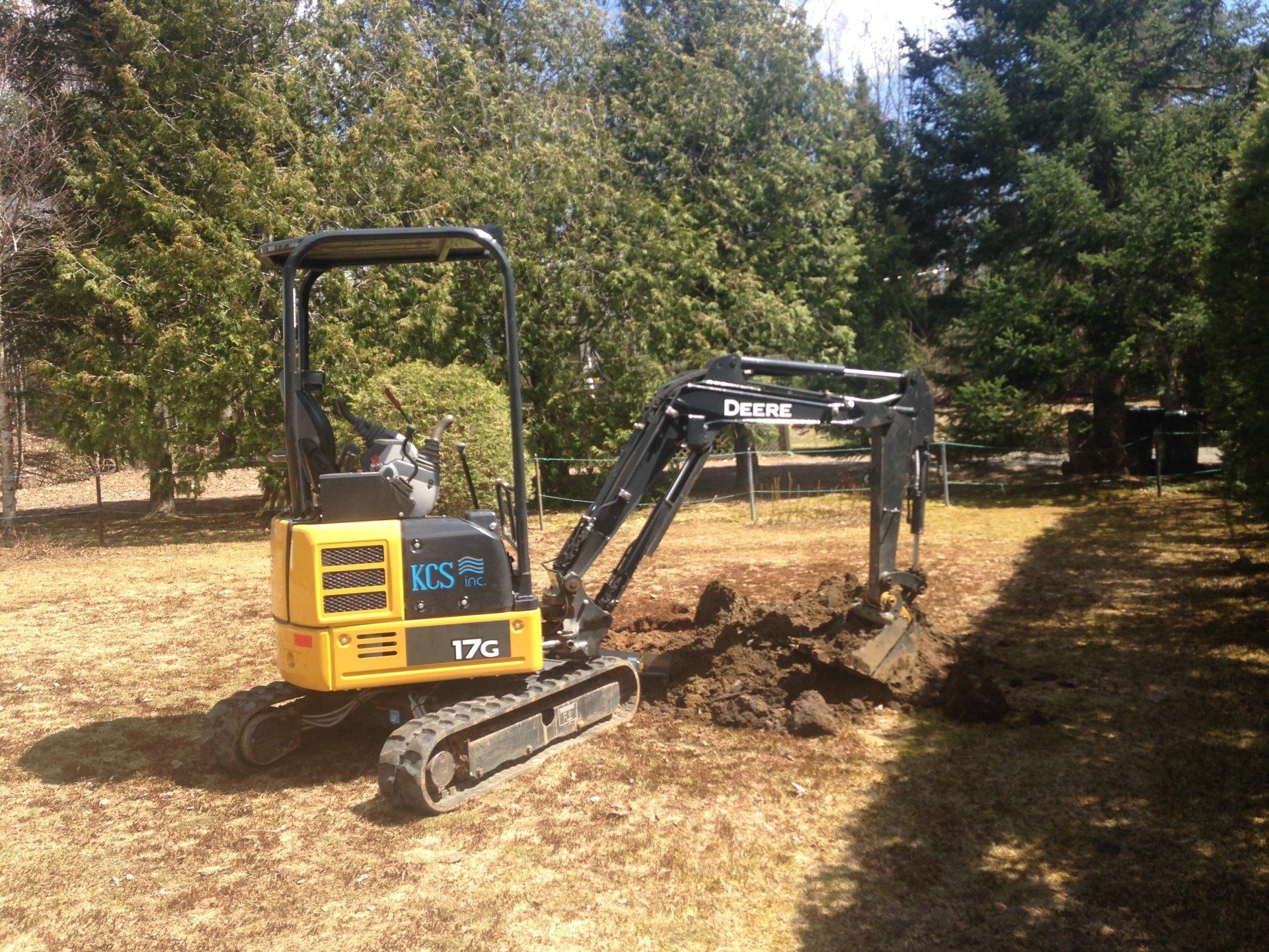 Yellow mini excavator digging in a grassy yard, near trees.