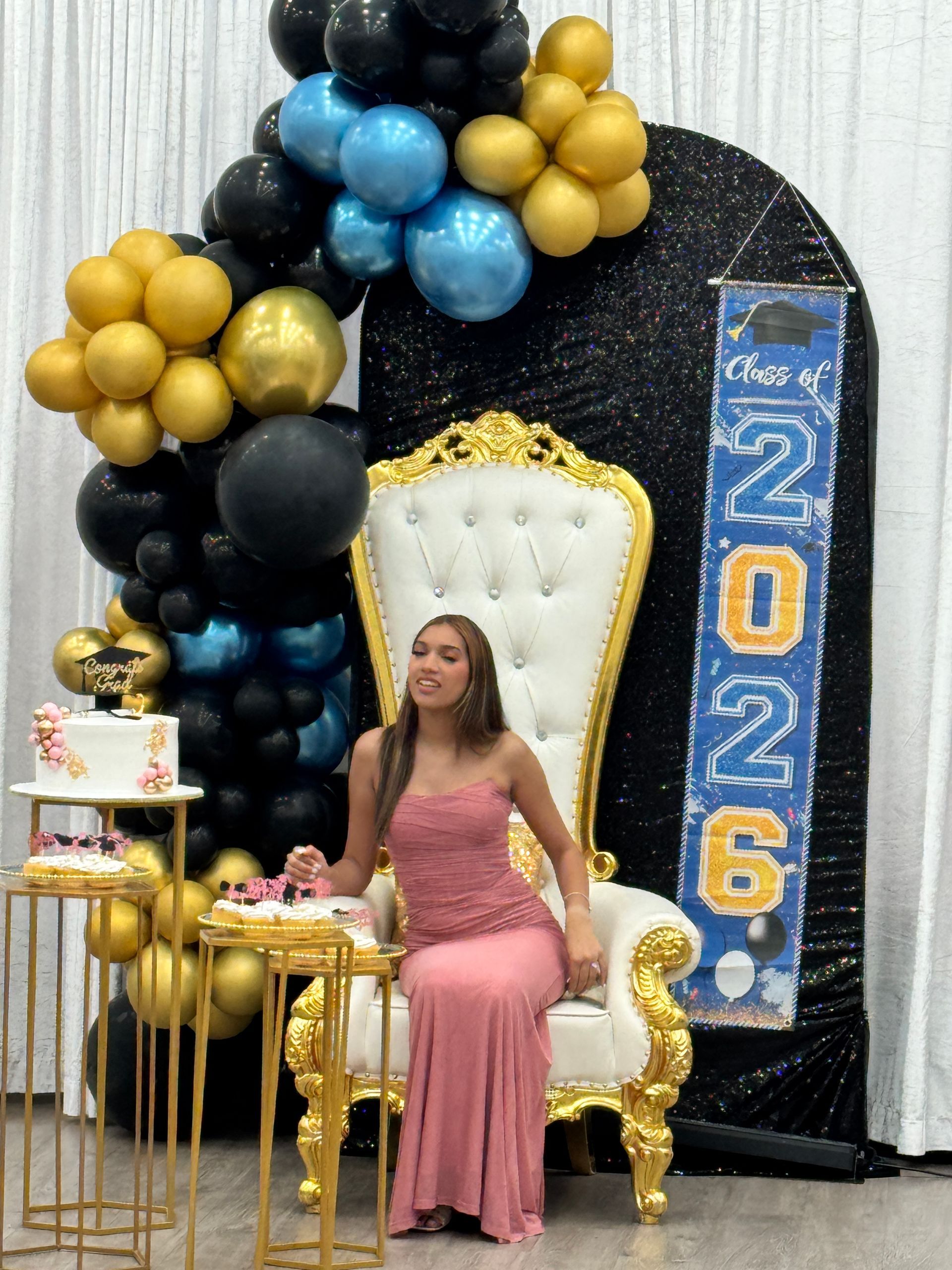 Woman seated on a gold throne at a 2026 celebration backdrop with black, gold, and blue balloons