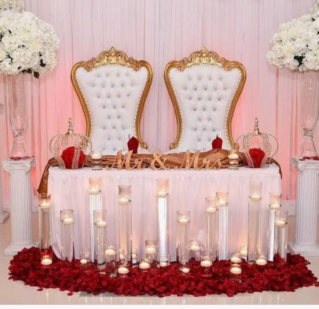 Elegant wedding stage with two white and gold chairs, pink drapes, candles, and red rose petals