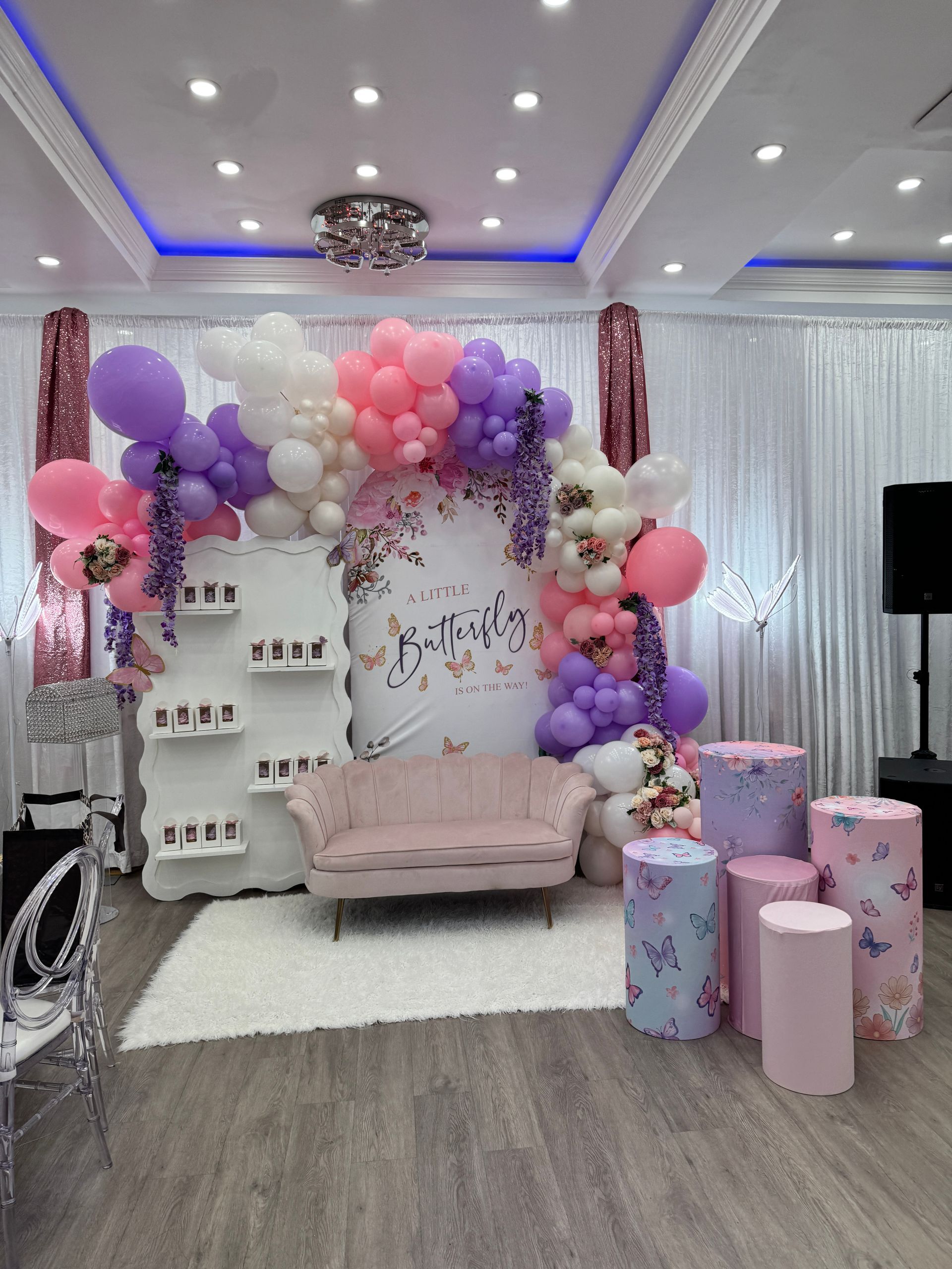 Pastel balloon-decorated celebration setup with pink couch, floral backdrop, and gift boxes in a living room