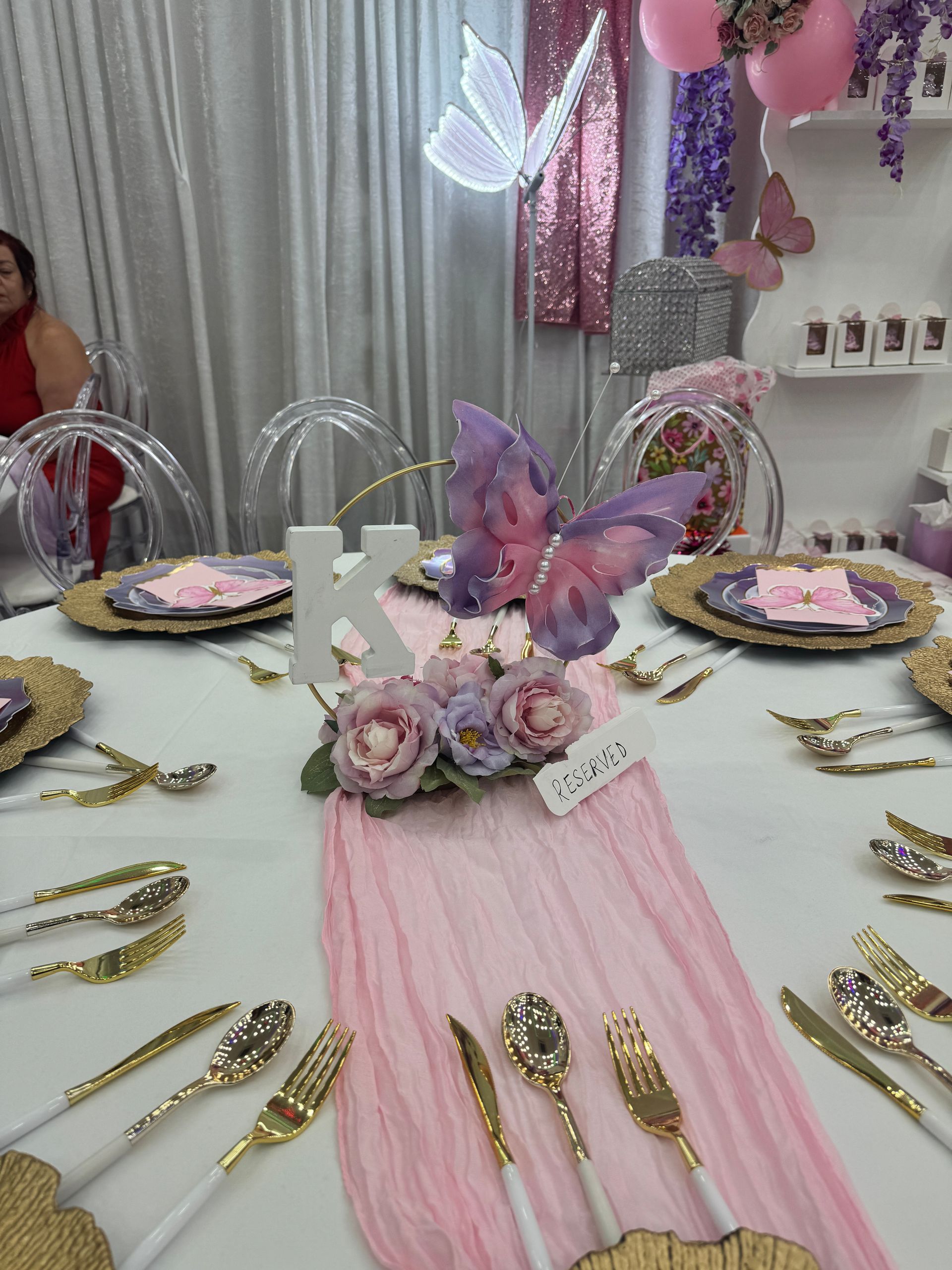 Pink butterfly-themed birthday table with gold utensils, flowers, and a decorated letter K centerpiece
