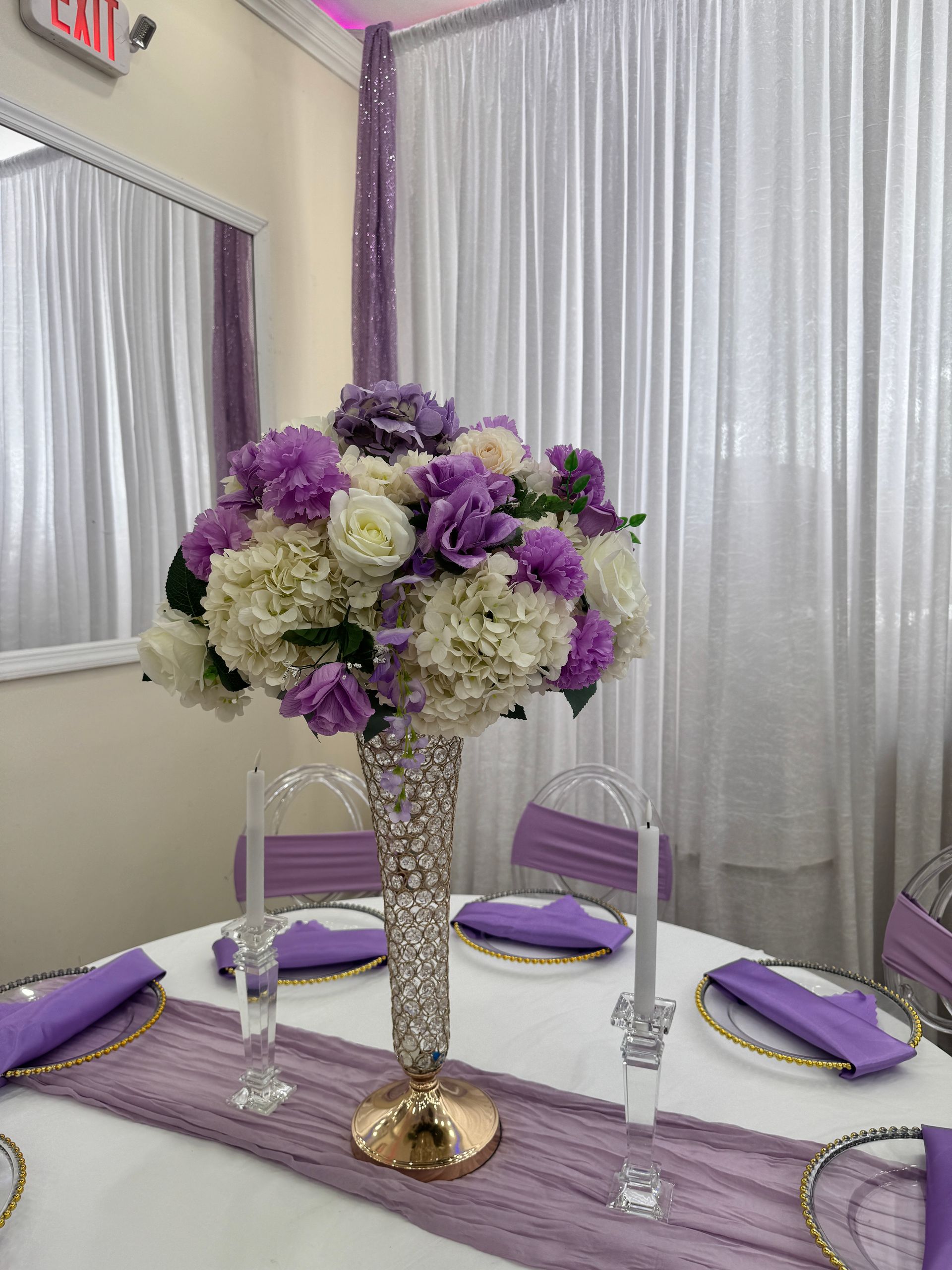 Elegant purple and white floral centerpiece on a decorated table with lavender chairs and silver drapes