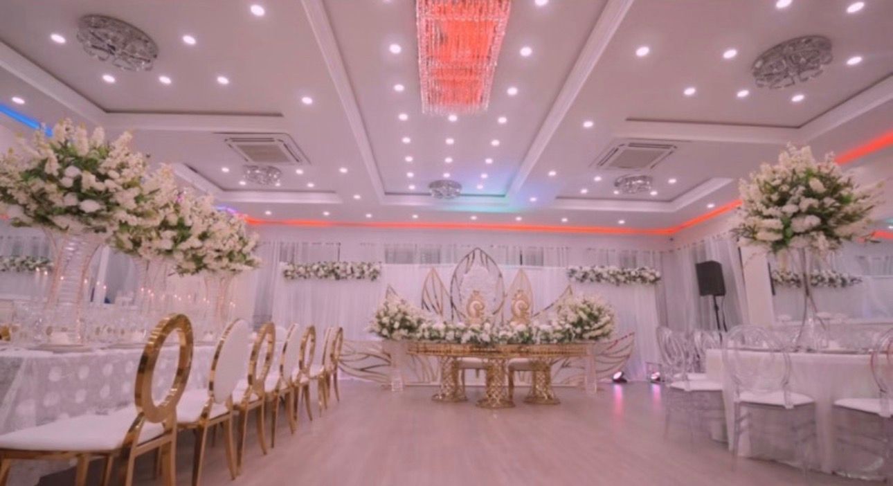 Elegant wedding reception hall with white and gold floral decor, round tables, and a central stage under pink lighting