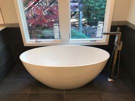 Modern white freestanding bathtub beside a window with outdoor view and dark floor tiles.