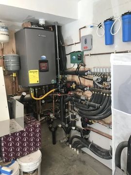 Utility room with a boiler, pipes, valves, and blue water filters on a white wall.