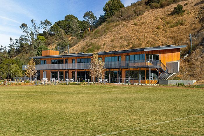 Modern two-story wooden building on a grassy field beside a wooded hillside