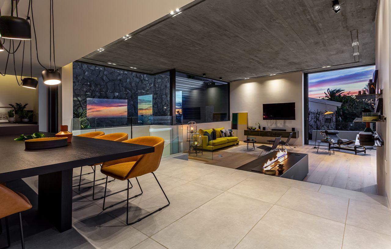 Modern open-plan apartment with kitchen, dining area, living room, and city sunset view through large windows