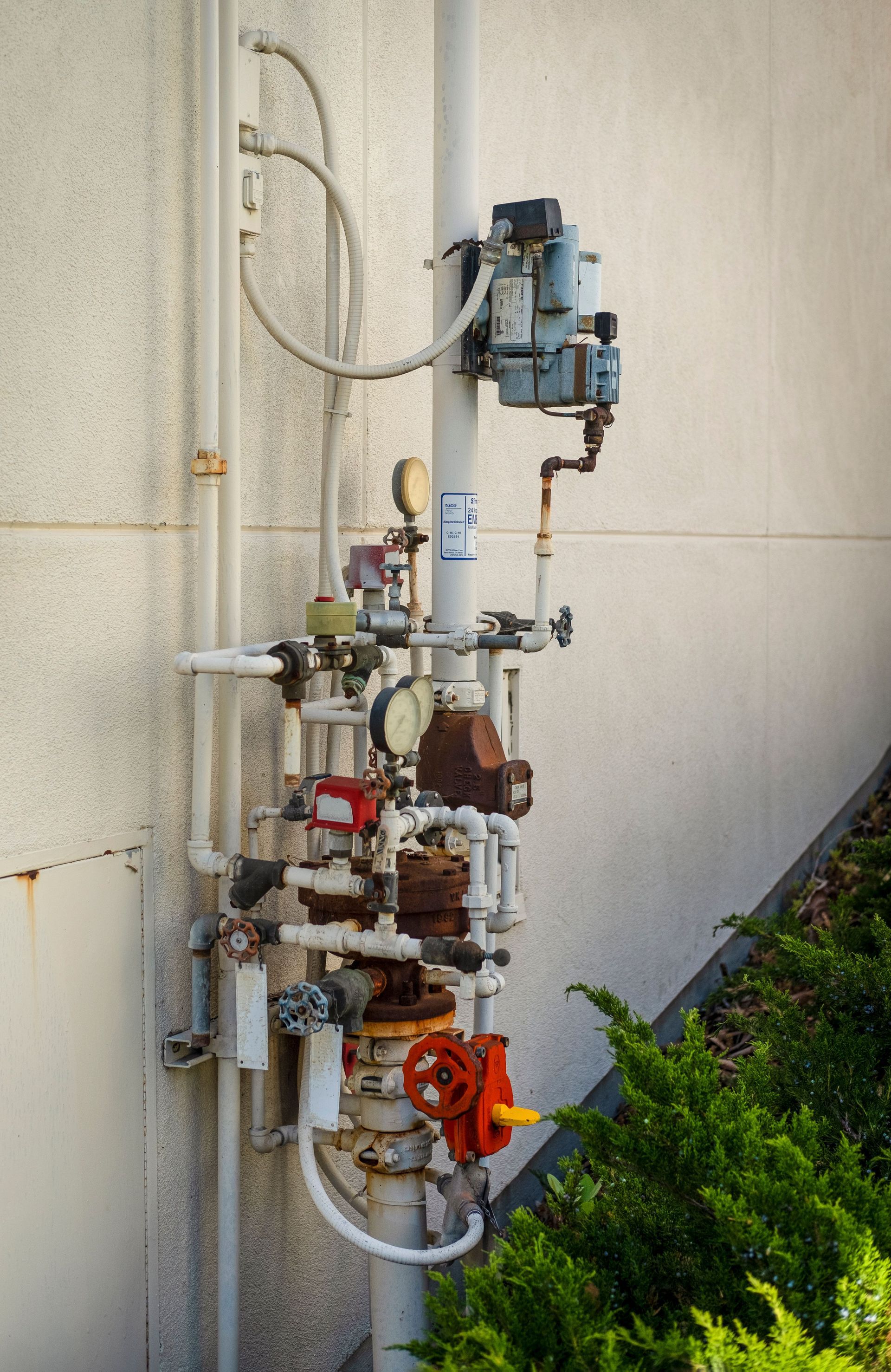 Outdoor industrial pipe and valve assembly mounted on a wall, with red, blue, and silver components.