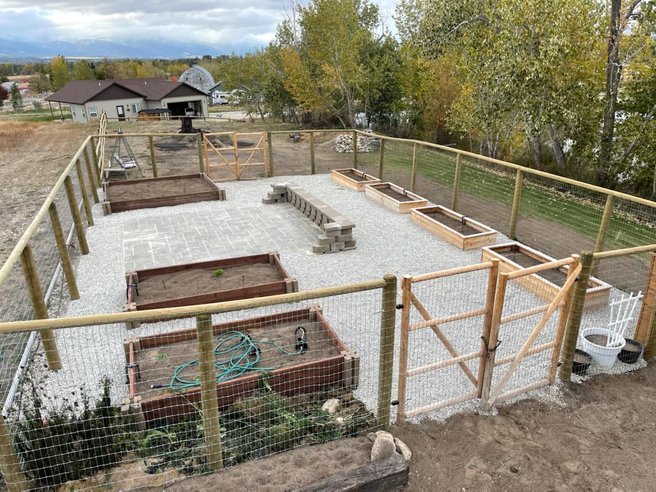 Fenced garden with raised beds and gravel. Wooden gates, hose, and a bench.