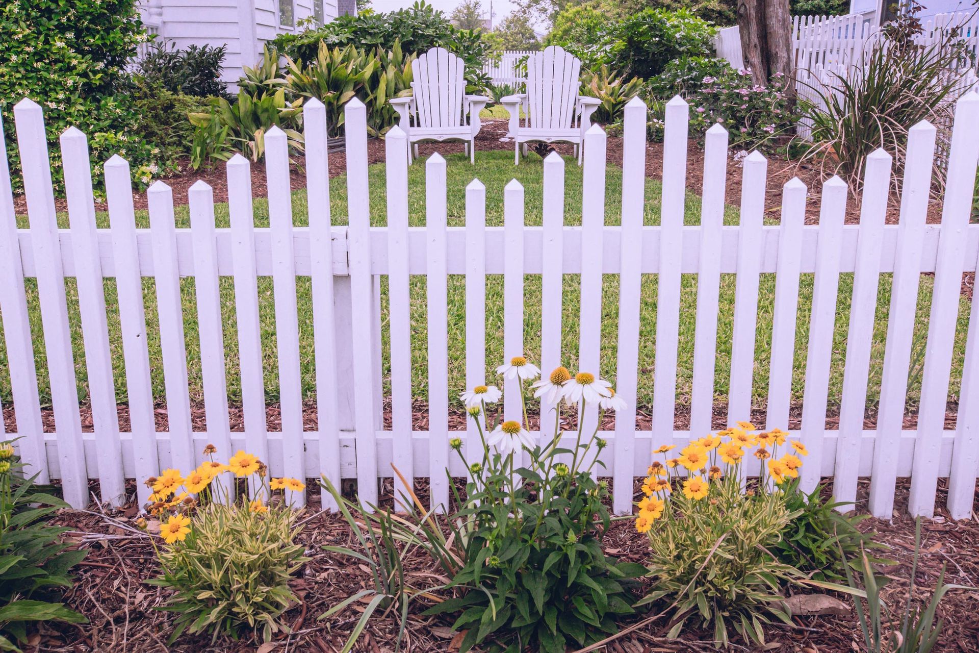 Man Screws Metal Fence Panel — Clifton, NJ — All Jersey Fence Co. A white picket fence surrounds a garden with yellow flowers.
