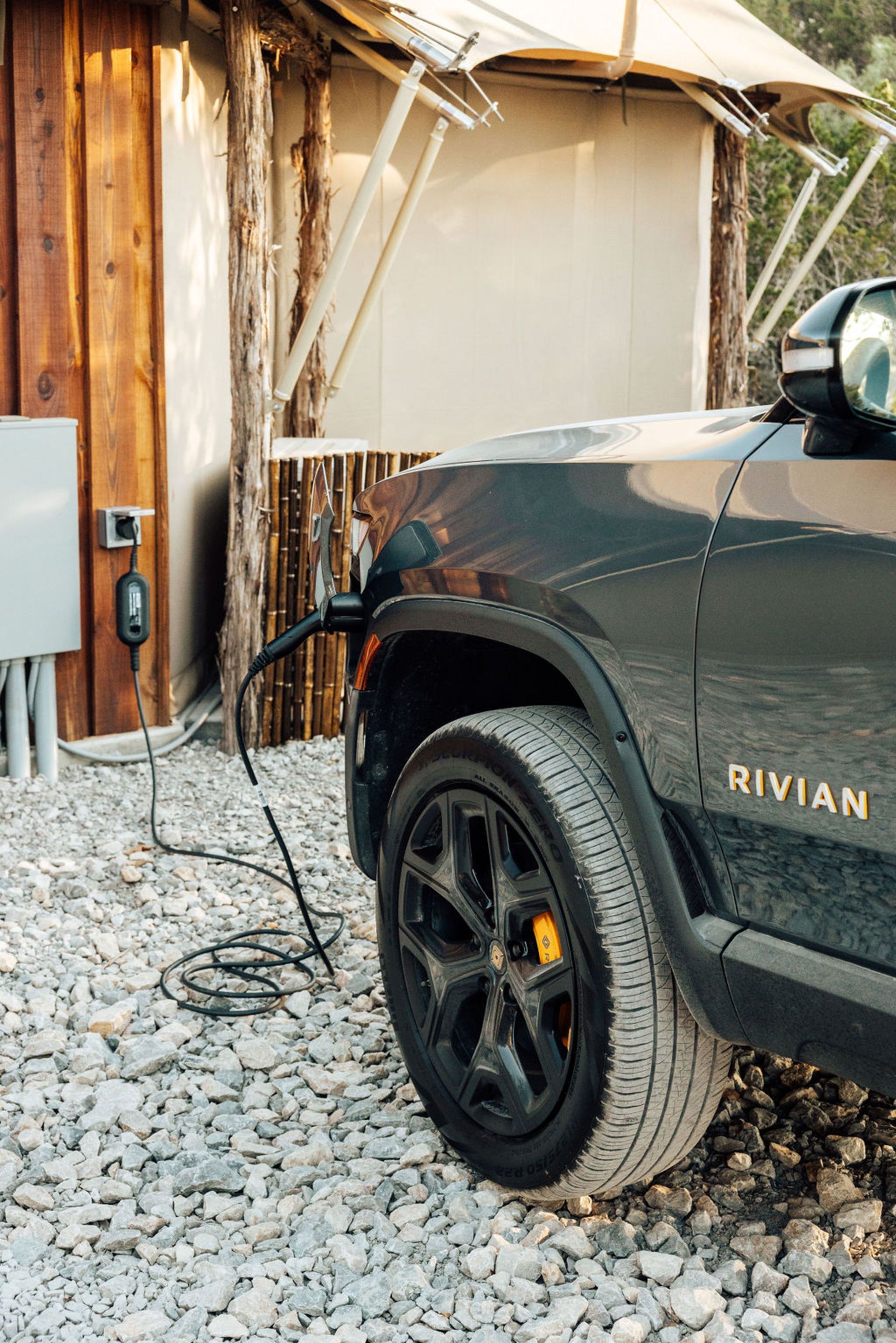 Dark Rivian truck charging near a wooden structure. Black wheels with orange calipers, gravel ground.