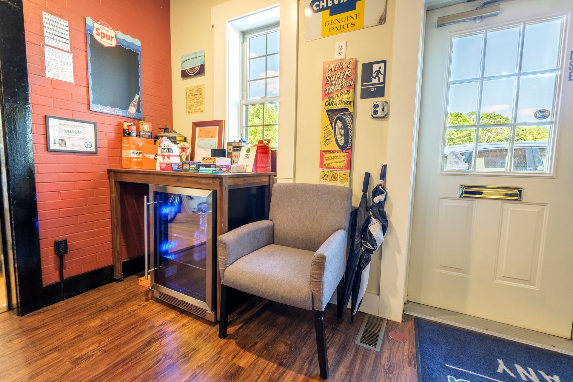 Interior with a small desk, mini-fridge, chair, and door. Red brick wall, brown wood floor. | Boalsburg Car Co.