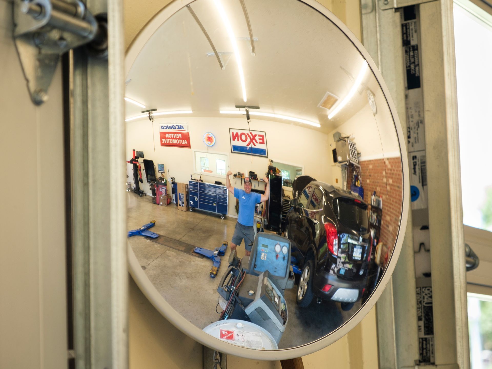 Circular mirror reflecting a cluttered auto repair shop with a person working on a vehicle. | Boalsburg Car Co.
