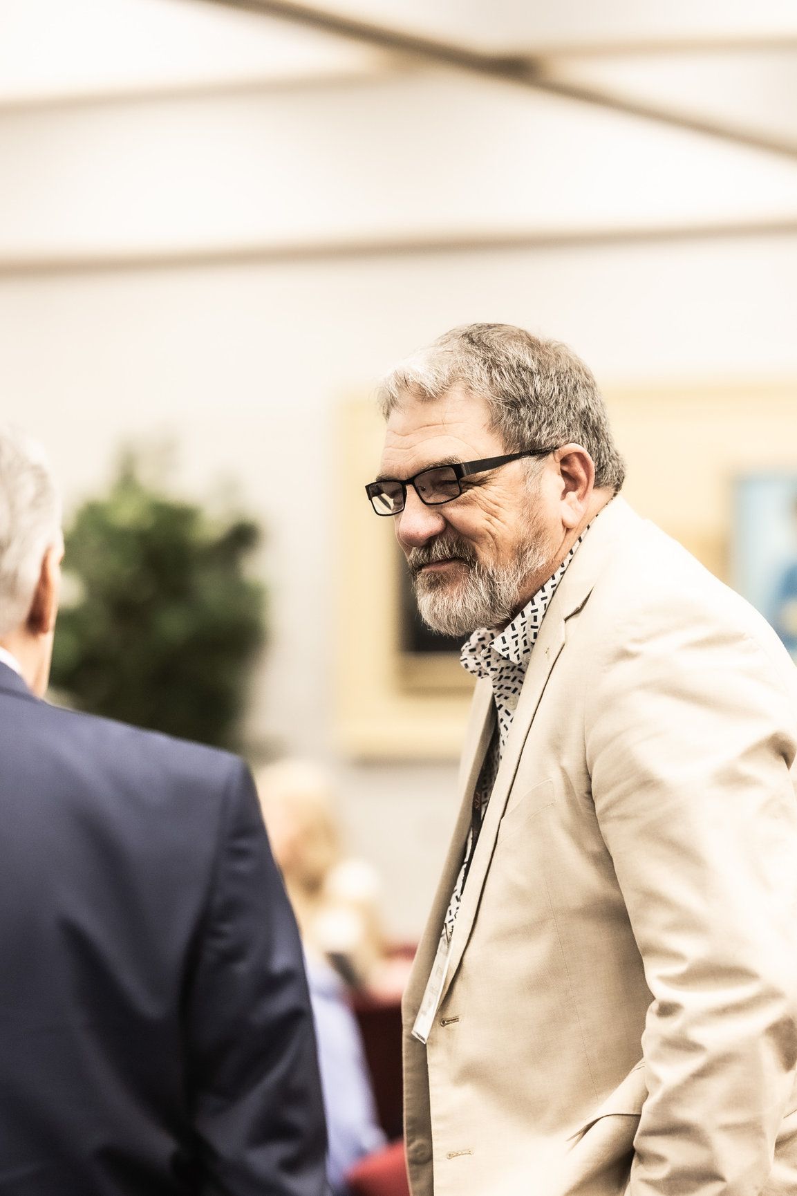 A man with glasses and a beard is standing next to another man in a suit.