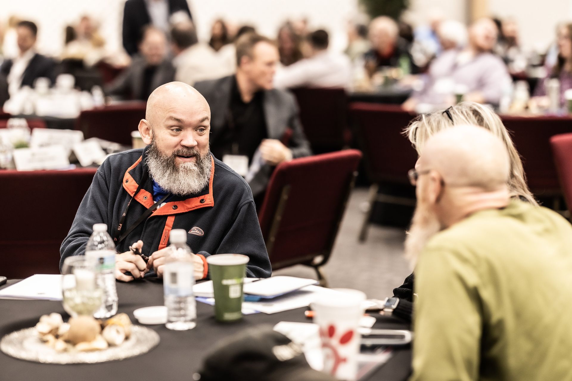 A man with a beard is sitting at a table talking to another man.